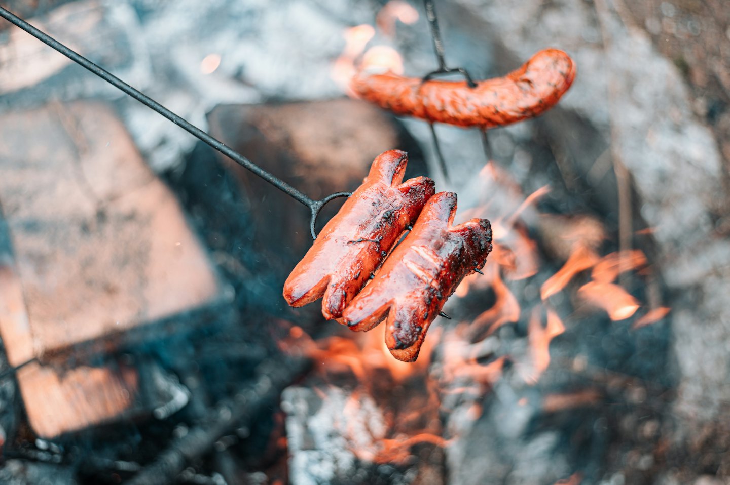 Sausages on metal skewers roast over an open campfire.